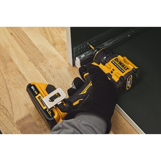 A person wearing a black glove is using a DEWALT DCD803NT cordless power drill to fasten a screw into a metal bracket inside a cabinet, on a wooden floor.