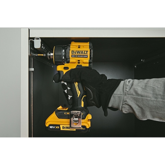 A person wearing a black glove is using a yellow and black DEWALT 18V XR brushless cordless drill to screw into a tight space inside a cabinet.
