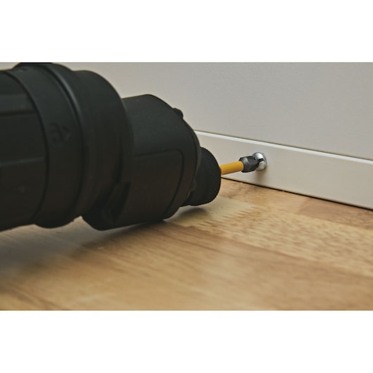 A close-up view of a black cordless drill driving a screw into a white baseboard situated above a wooden floor.