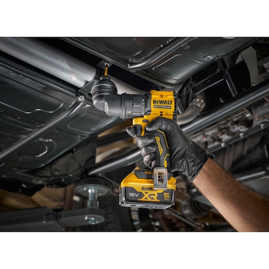 A gloved hand holds a yellow DEWALT 18V XR cordless drill, driving a screw into metal piping under a vehicle chassis.