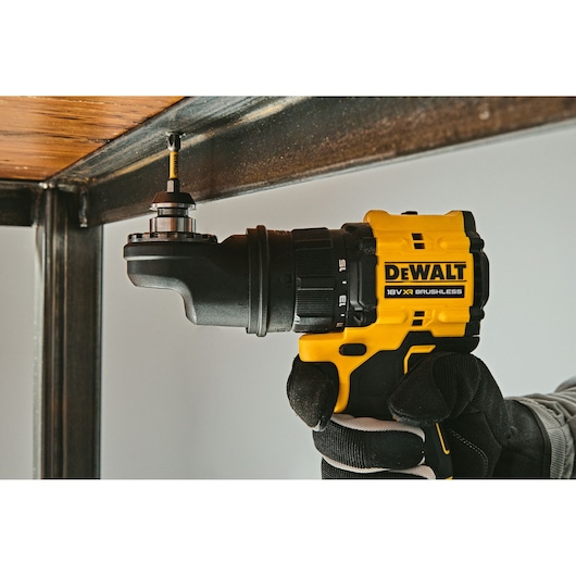 A close-up of a person using a yellow and black DEWALT 18V XR brushless drill, fitted with an angled attachment, to fasten a screw under a metal-framed wooden surface.