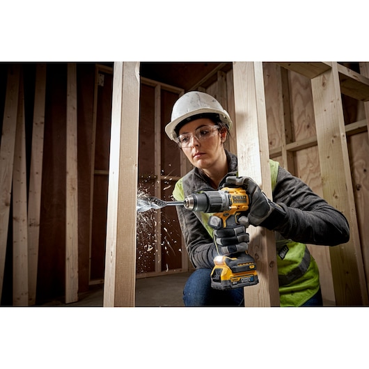 A construction worker wearing a safety helmet and vest is using a DEWALT cordless drill (SKU: DCD805E1T) to drill into a wooden beam, causing wood chips to fly. The drill is yellow and black and used in a framing environment.