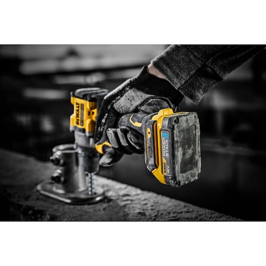 A gloved hand holds a DEWALT cordless power drill with a POWERSTACK 18V battery, drilling into a metal surface in an industrial setting.