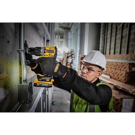 A person wearing protective gear and a safety helmet uses a DEWALT DCD805E2T cordless power drill to work on a metal pipe attached to a wall inside an industrial or construction site.