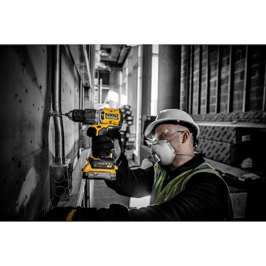 A worker wearing a safety helmet and gloves is using a yellow DEWALT DCD805E2T cordless drill to fasten a metal pipe in an industrial construction setting.