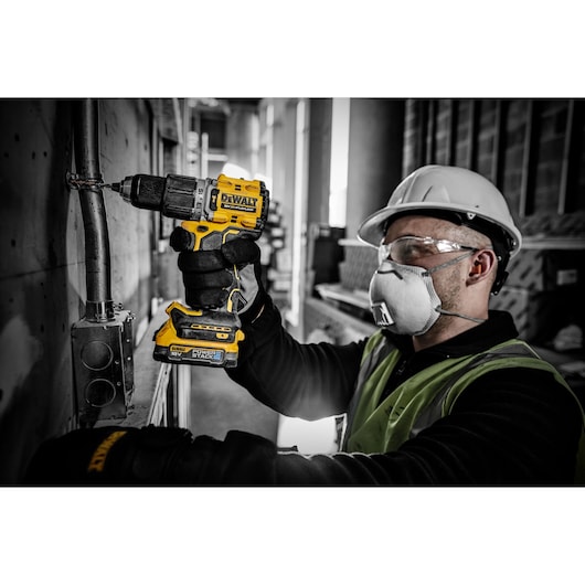 A person wearing a safety helmet and high-visibility vest is using a DEWALT DCD805E2T cordless drill to work on a metal pipe attached to a wall in an industrial setting. The person's face is blurred for privacy.