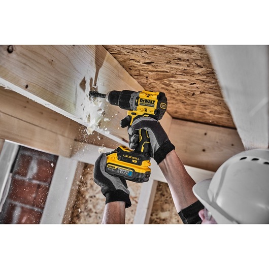 Person uses DEWALT cordless drill to bore into wood ceiling. Wood chips scatter. Worker wears gloves and helmet.