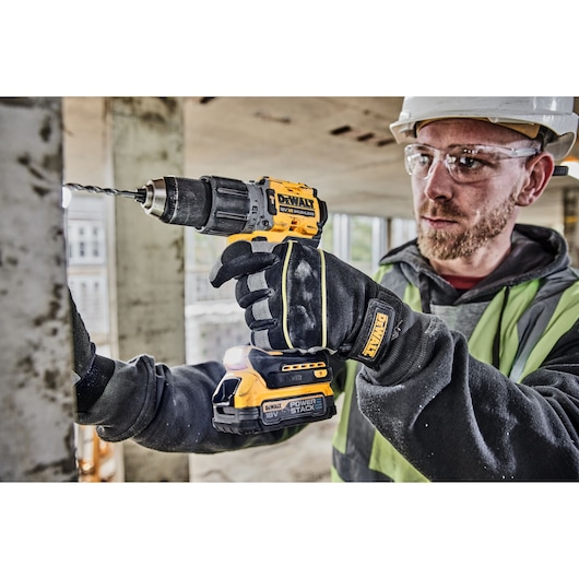 A person wearing safety gloves and a safety vest is using a DEWALT 18V cordless drill to drill into a metal surface at a construction site. The drill is branded DEWALT and fitted with a POWERSTACK battery.