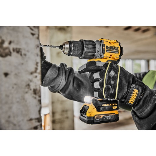 A person wearing black work gloves is using a yellow and black DEWALT cordless drill to drill into a concrete surface indoors.