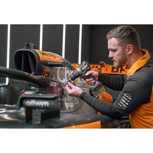 A person using a DEWALT DCD85MM1T cordless drill on the side of an orange and black race car in a workshop. The person's face is blurred for privacy, and the car has visible sponsor logos.