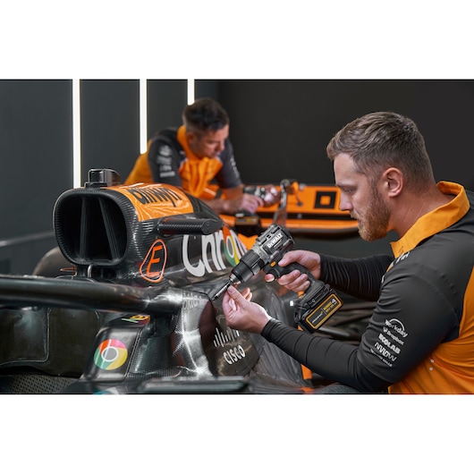 Two people in orange and black uniforms work on a Formula 1 race car. One person in the foreground uses a DEWALT cordless drill on the car's side. The car displays various sponsor logos, and both individuals' faces are blurred.