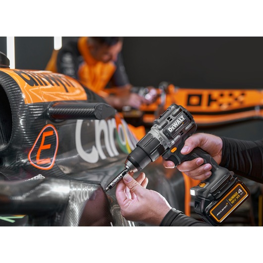 A person is using a DEWALT DCD85MM1T cordless drill to work on a carbon fiber body panel of a race car. The car features orange and black livery with visible sponsorship logos. The focus is on the drill and the hands performing maintenance or assembly, with another person blurred in the background.