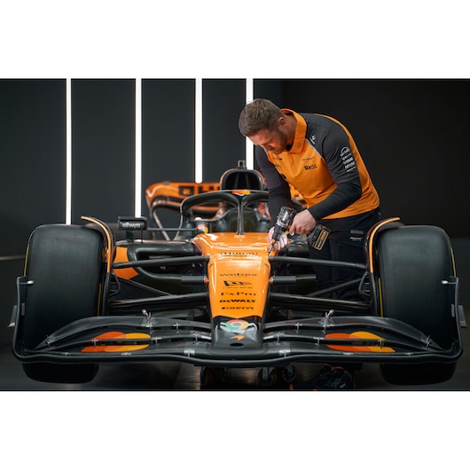 A person wearing an orange and black DEWALT branded uniform uses a DEWALT power tool on the front section of an orange Formula 1 race car in a modern workshop. The person's face is blurred.