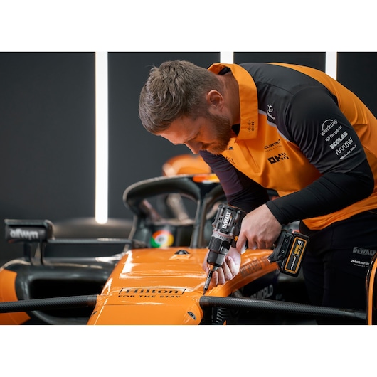 A person wearing an orange and black uniform uses a DEWALT cordless drill to work on an orange race car, focusing on the body near the front. The person's face is blurred.