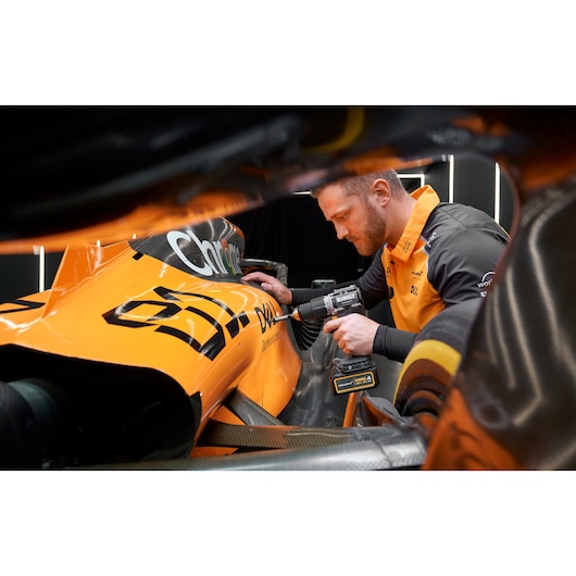 A person is using a DEWALT cordless drill to work on the body of a bright orange race car, with the car's branding visible. The individual's face is blurred for privacy.