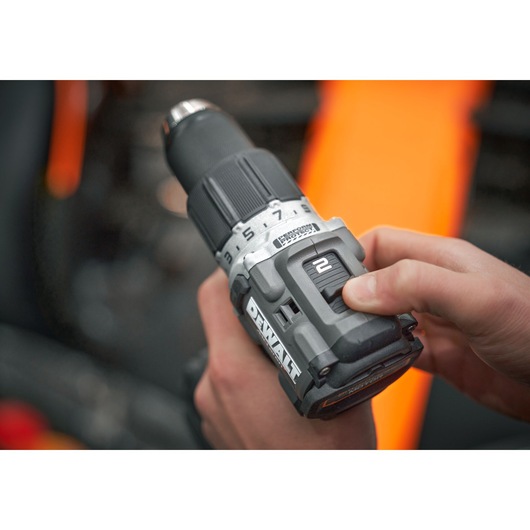 A close-up view of a person holding a DEWALT cordless drill, with their thumb adjusting the speed setting switch. The background is blurred, showing some orange and dark colors.
