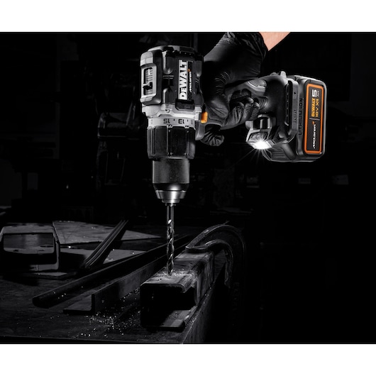 A DEWALT cordless drill being used to drill into a metal surface, held by a gloved hand in a workshop setting. The drill has a visible LED light and a 5 Ah battery attached.