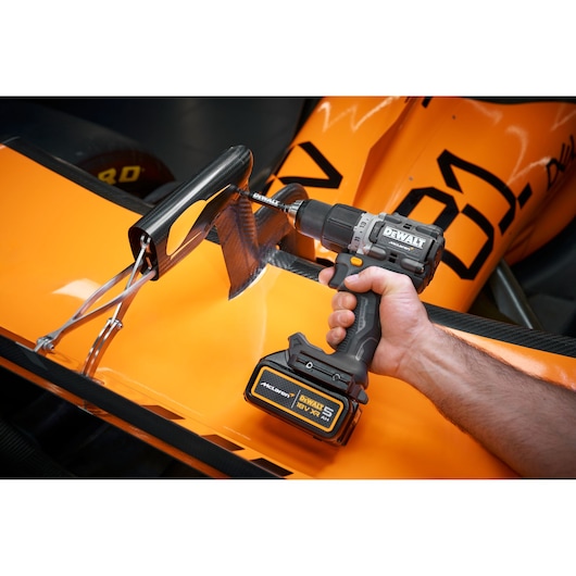 A person uses a DEWALT cordless drill to work on a carbon fiber part attached to an orange race car.