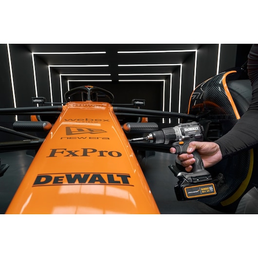 A person uses a DEWALT cordless drill on an orange Formula 1 race car, featuring prominent sponsor logos including DEWALT, FxPro, New Era, and Webex. The scene is set in a modern, illuminated garage.