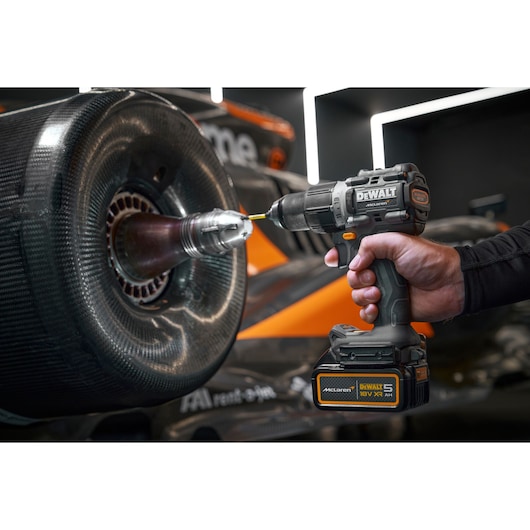 A close-up of a DEWALT cordless drill being used on a race car wheel, with a hand holding the tool. The wheel has a carbon fiber finish and the car features orange and black colors.