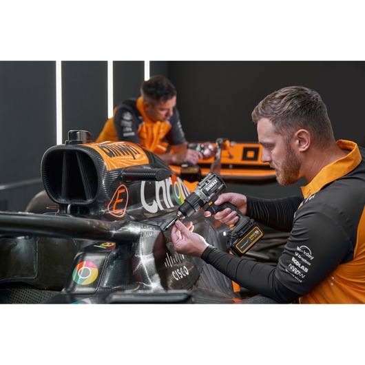 Two individuals wearing orange and black uniforms are working on a race car. One is using a DEWALT cordless power drill, model DCD85MP2T, on the car's body. The individuals' faces are blurred for privacy.