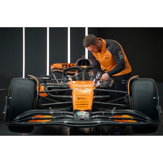 A person in an orange and black DEWALT uniform uses a power tool to work on the front of an orange and black Formula 1 racing car in a garage setting.
