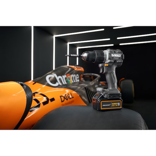 A DEWALT cordless power drill is placed on the tire of an orange race car, with illuminated white lines in the background. The car features branding such as 'Chrome' and 'DELL Technologies.'