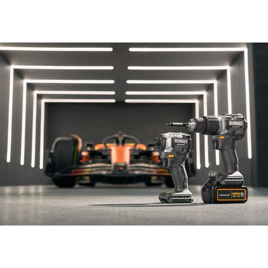 Two DEWALT power tools, including a drill and an impact driver, are positioned in the foreground of a modern, illuminated garage with a racing car in the background.