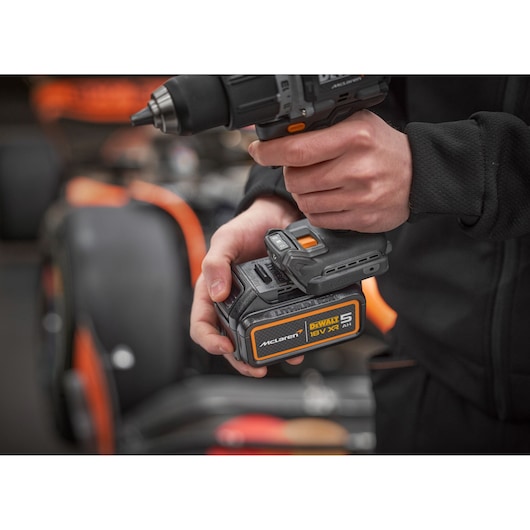 A close-up of a person holding a DEWALT cordless drill with an 18V XR 5Ah McLaren edition battery, with the background showing an out-of-focus vehicle.