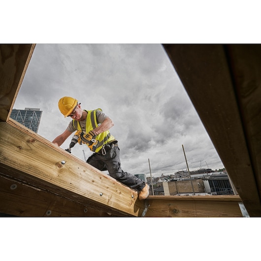 A construction worker wearing a yellow hard hat and high-visibility vest uses a DEWALT cordless drill while working on a wooden beam outdoors, with buildings visible in the background under a cloudy sky.