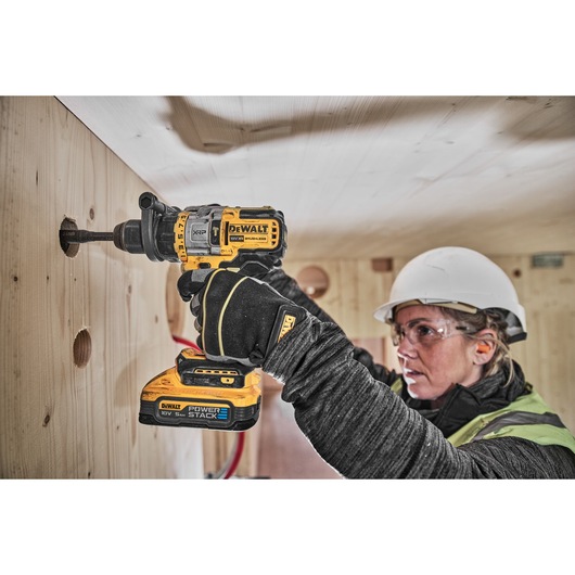 A person wearing safety gear is using a DEWALT DCD999H2T cordless drill to bore a hole into a wooden wall. The drill is fitted with a POWERSTACK battery.