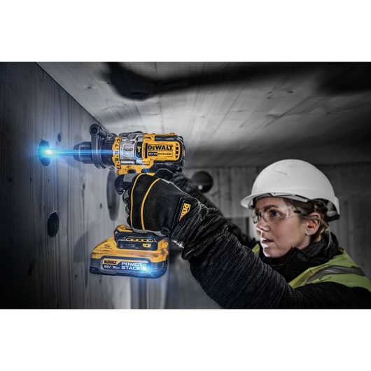 A person wearing safety gear uses a DEWALT DCD999H2T PowerStack cordless drill to bore a hole into a wooden wall. The drill is illuminated, and the person's face is blurred for privacy.