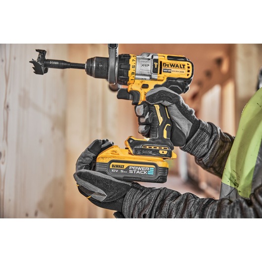 A close-up of a gloved worker holding a DEWALT DCD999H2T cordless drill with a POWERSTACK 18V 5Ah battery, drilling into wood.