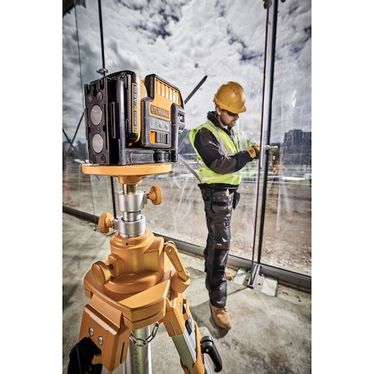 A DEWALT laser level (SKU: DCE0822D1R) mounted on a tripod stands in the foreground of a construction site. A worker in a high-visibility vest and helmet is seen in the background using tools near large glass panels.