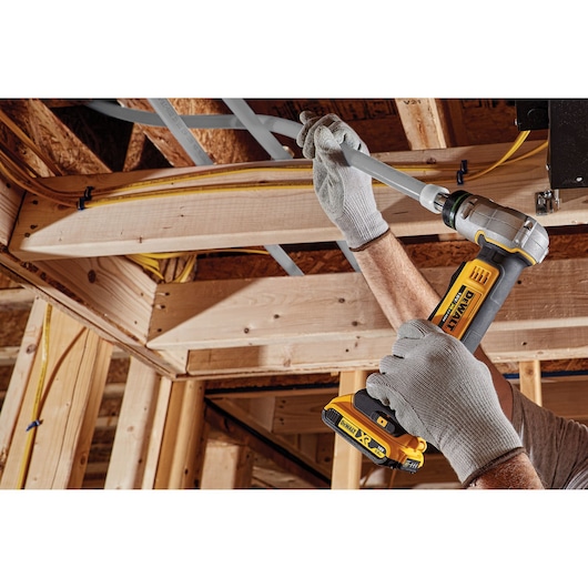 A person wearing gray work gloves uses a DEWALT cordless pipe crimping tool to secure a gray plastic pipe to wooden framing during construction.