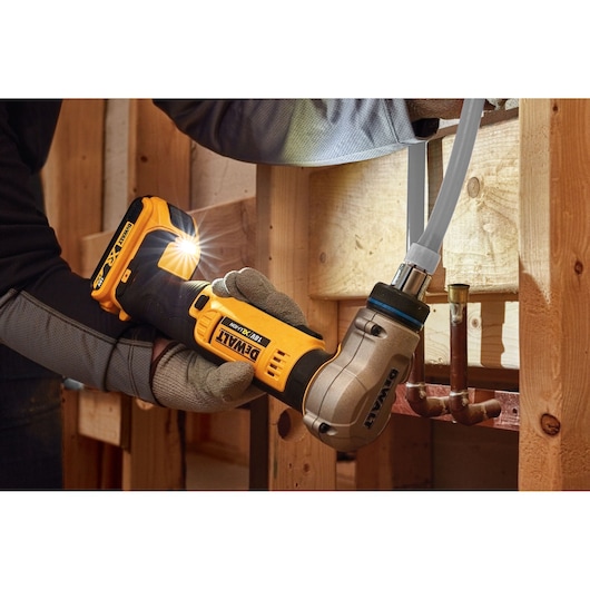 A person wearing work gloves uses a DEWALT DCE400D2 18V cordless pipe crimping tool to connect a pipe inside a wooden framed construction area.