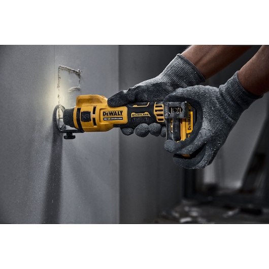 A person wearing grey gloves is using a DEWALT cordless rotary tool to cut a square hole in a drywall panel.