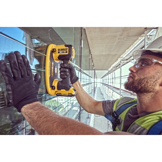 A person wearing gloves and a safety vest is using a DEWALT DCE590 18V XR cordless tool against a glass surface on a construction site. The face is blurred for privacy.