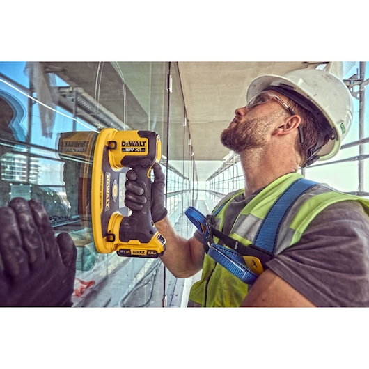 A construction worker wearing a safety helmet and high-visibility vest is using a DEWALT DCE590 18V XR handheld tool to install or work on a glass panel, with a reflection visible on the glass surface.
