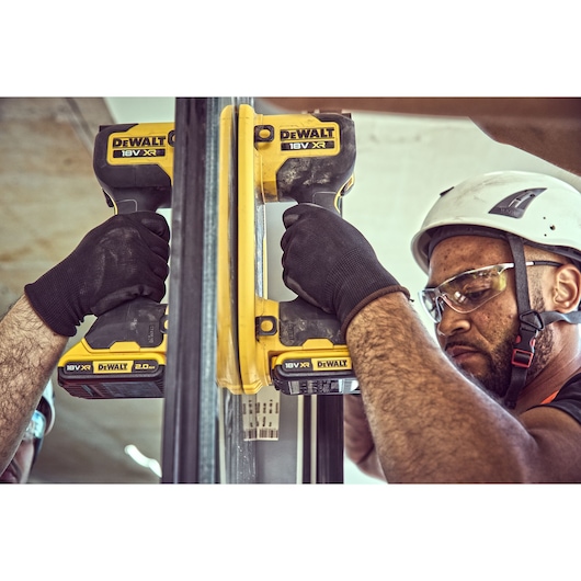 Two people wearing gloves and safety gear use DEWALT 18V XR cordless tools to handle a large sheet of glass. The tools are pressed firmly to the glass surface.