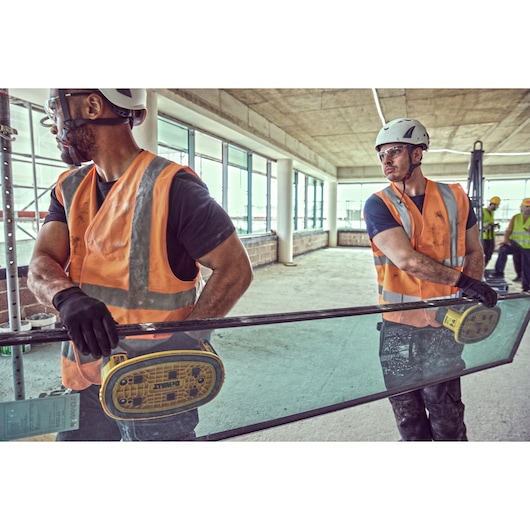 Two construction workers wearing safety helmets, gloves, and orange reflective vests are using DEWALT DCE590 tools to lift and carry a large glass panel at an indoor construction site. Other workers and construction materials are visible in the background.