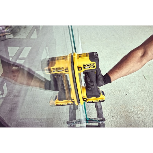 A person using a yellow DEWALT power tool with a suction attachment to handle or move a large glass sheet. The tool is labeled with 18V XR and is held by a gloved hand.