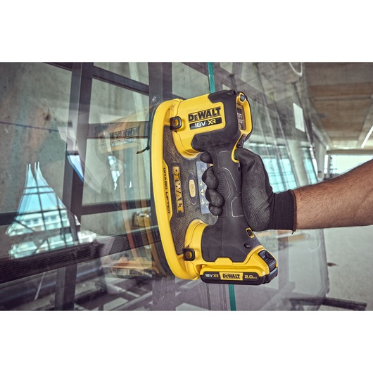 A DEWALT 18V XR cordless glass lifter tool is being used to handle a large glass pane. A gloved hand grips the yellow and black tool, which is pressed against the glass in a modern industrial setting.