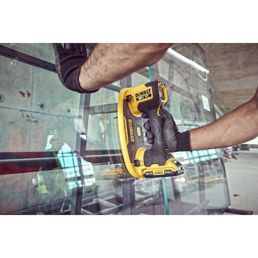 A person wearing black gloves is using a DEWALT DCE590 cordless tool to handle a sheet of glass in an industrial setting.