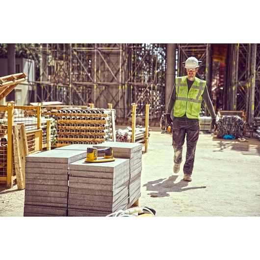 A DEWALT power tool (DCE590) is placed on stacked concrete slabs at a construction site, with a worker wearing a safety vest and helmet walking nearby. The background shows building materials and scaffolding.