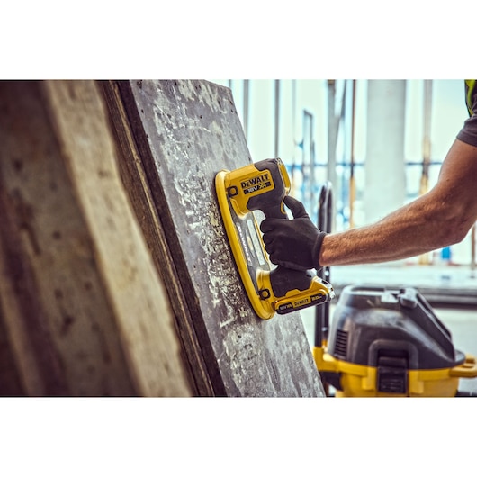 A person wearing a black glove uses a DEWALT 18V XR cordless tool on a vertical concrete surface, likely for chiseling or breaking, with a shop vacuum visible in the background.