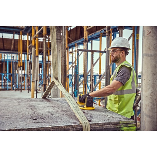 A construction worker wearing a reflective vest and hard hat operates a DEWALT DCE590 tool on a large concrete slab, secured with heavy-duty straps, at a building site with scaffolding and structural columns.