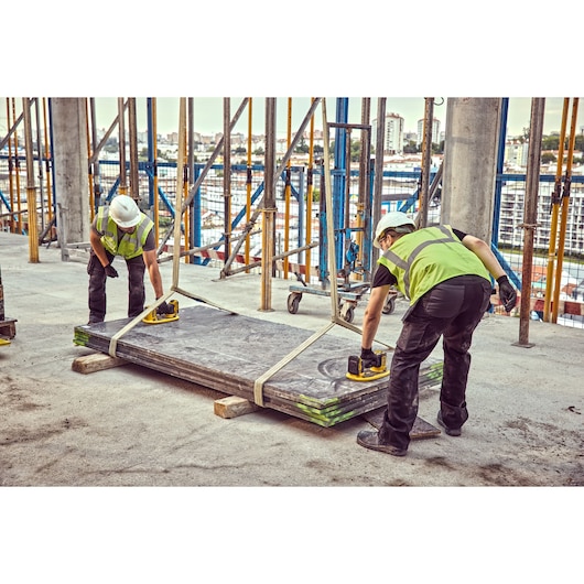 Two workers wearing safety vests and helmets are using DEWALT power tools to handle large metal sheets on a construction site. The sheets are secured with straps and surrounded by scaffolding.