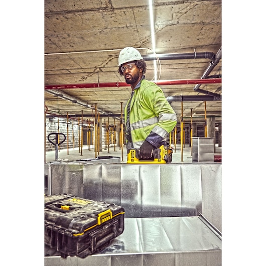 A construction worker wearing a hi-vis jacket and helmet is holding a DEWALT DCE590 tool in an industrial environment, with metal ductwork and a DEWALT tool case in the foreground.