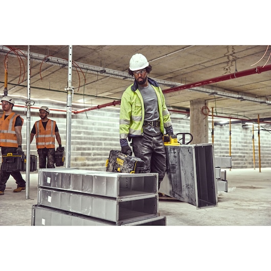 A construction worker wearing a reflective jacket and hard hat is carrying a DEWALT tool case and a large metal duct in an unfinished building. Two other workers in safety vests and helmets are in the background, also holding tool cases. The setting shows concrete floors, exposed pipes, metal scaffolding, and stacks of metal ducts.
